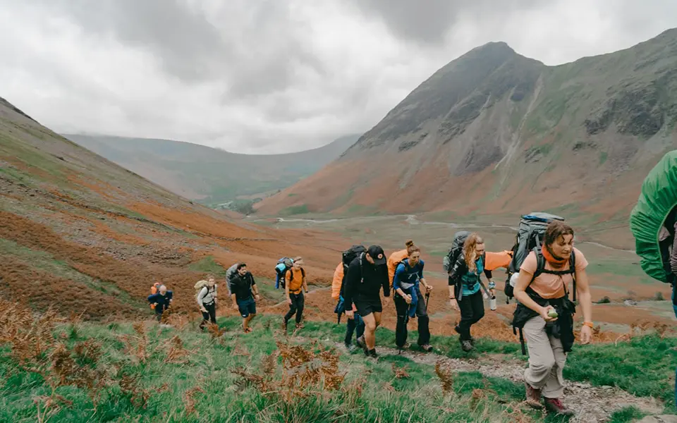 A group of hikers ascending a grassy hillside in a mountainous landscape, carrying backpacks and wearing outdoor gear. The scene is set against the dramatic backdrop of the Lake District, with cloudy skies and rugged peaks in the distance.