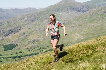 Lauren participating in a fell race, running along a grassy slope against the sunny backdrop of the rugged Lake District landscape.