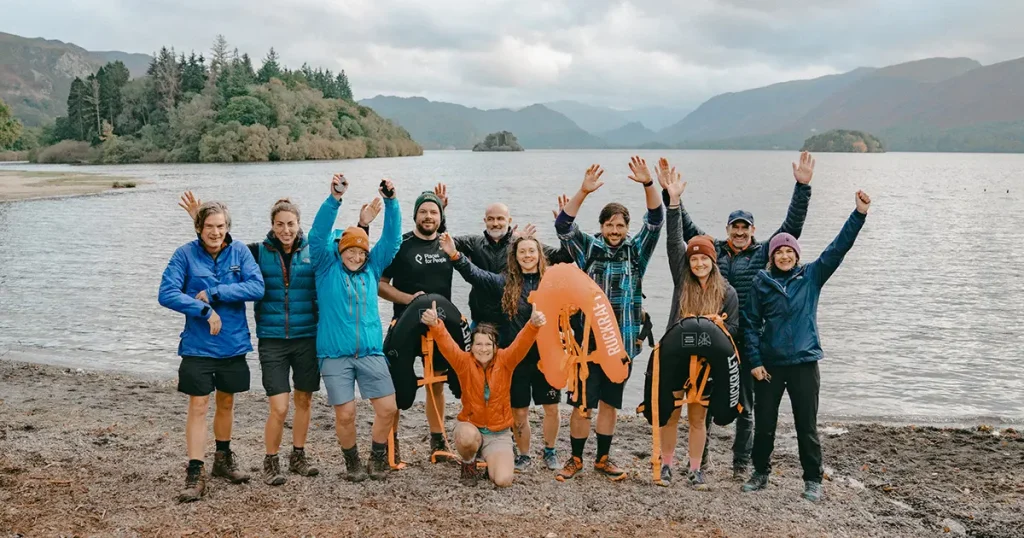 A group of adventurers joyfully celebrates the conclusion of their Ruck Raft experience, guided by Lauren Munro-Bennett, amidst the breathtaking scenery of the Lake District.