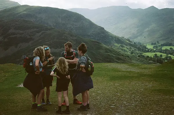 Lauren teaching a family of four navigation and route-finding techniques while standing atop a fell, surrounded by panoramic views of the Lake District.